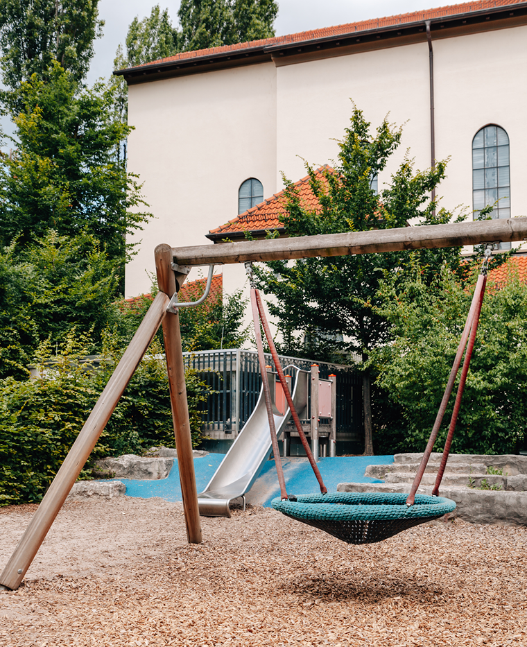 Garten einer Kita mit Vogelnestschaukel im Vordergrund und Rutsche im Hintergrund.