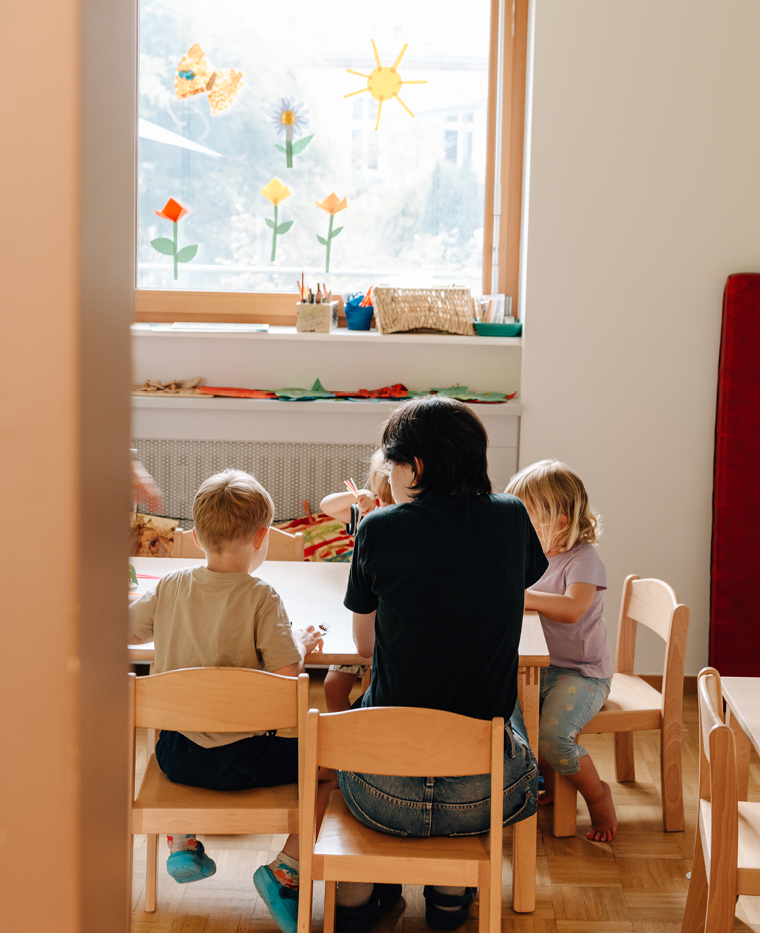 Gruppenraum mit Kindern und einer Mitarbeitenden, die am Tisch sitzen