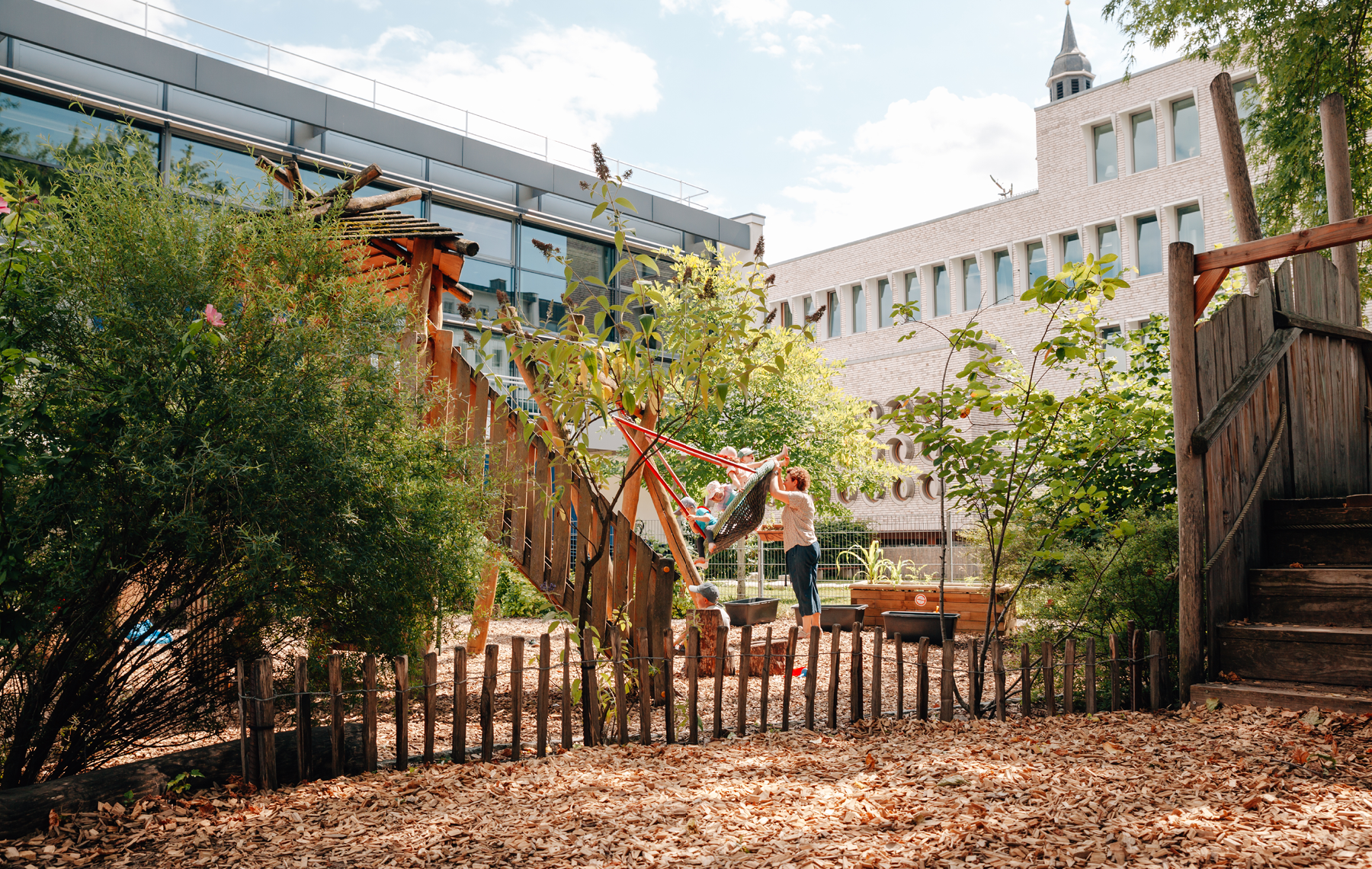 Garten der Kita in dem Kinder in der Vogelnestschaukel von einer Mitarbeiterin angeschubst werden